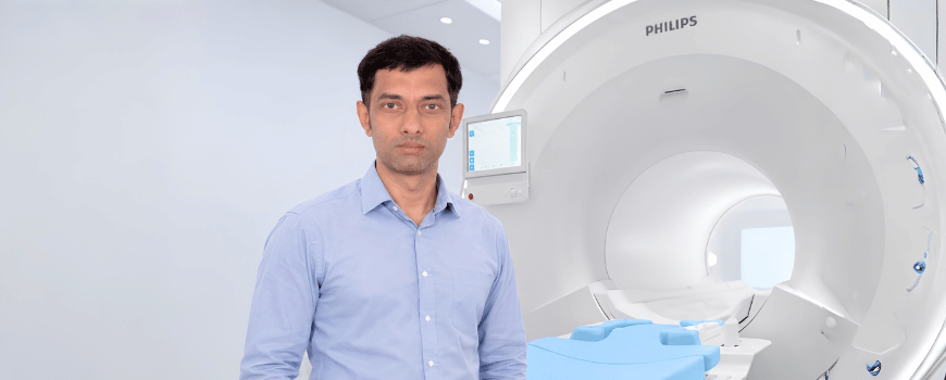 A calm male healthcare professional in a light blue shirt stands beside a large white Philips MRI scanner in a bright, modern medical imaging room.