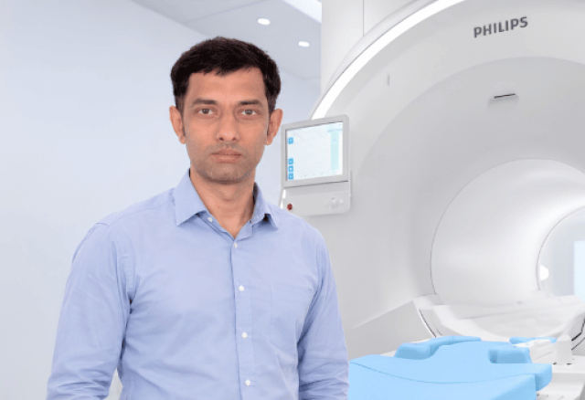 A calm male healthcare professional in a light blue shirt stands beside a large white Philips MRI scanner in a bright, modern medical imaging room.